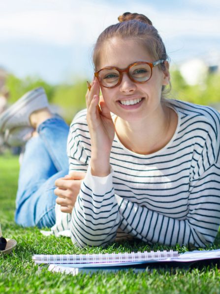 Summer outdoor photo of young happy European woman lying on green lawn in park dressed in striped top and jeans, wearing eyeglasses enjoying both leisure time and studying for classes outdoors